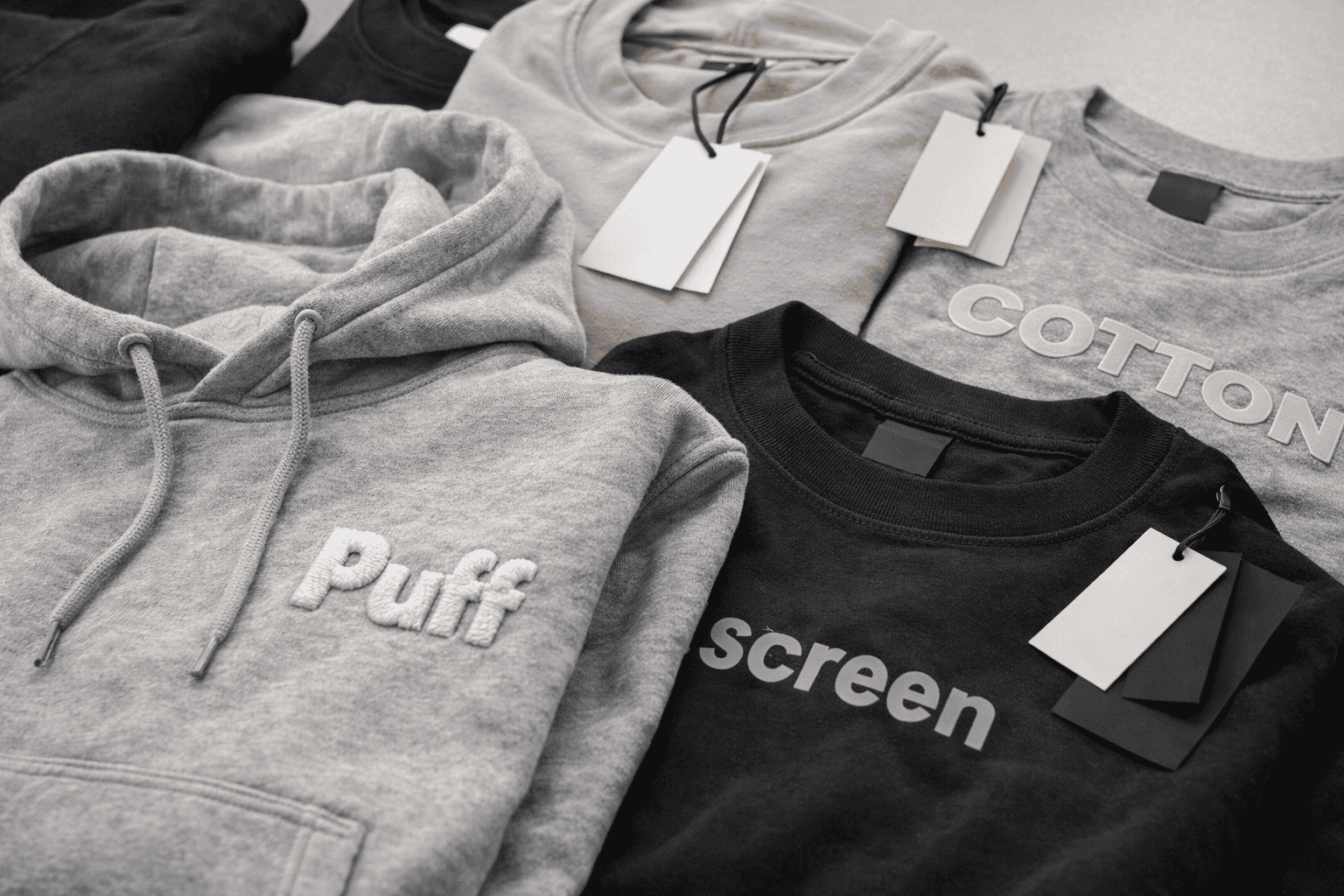 Close-up of custom streetwear hoodie and t-shirt showing embroidery, puff print, screen printing and silicone logo techniques, visible stitching and fabric texture, clean table setup, soft studio lighting, ultra realistic, subtle OHSURE branding --ar 16:9 --style raw --v 6