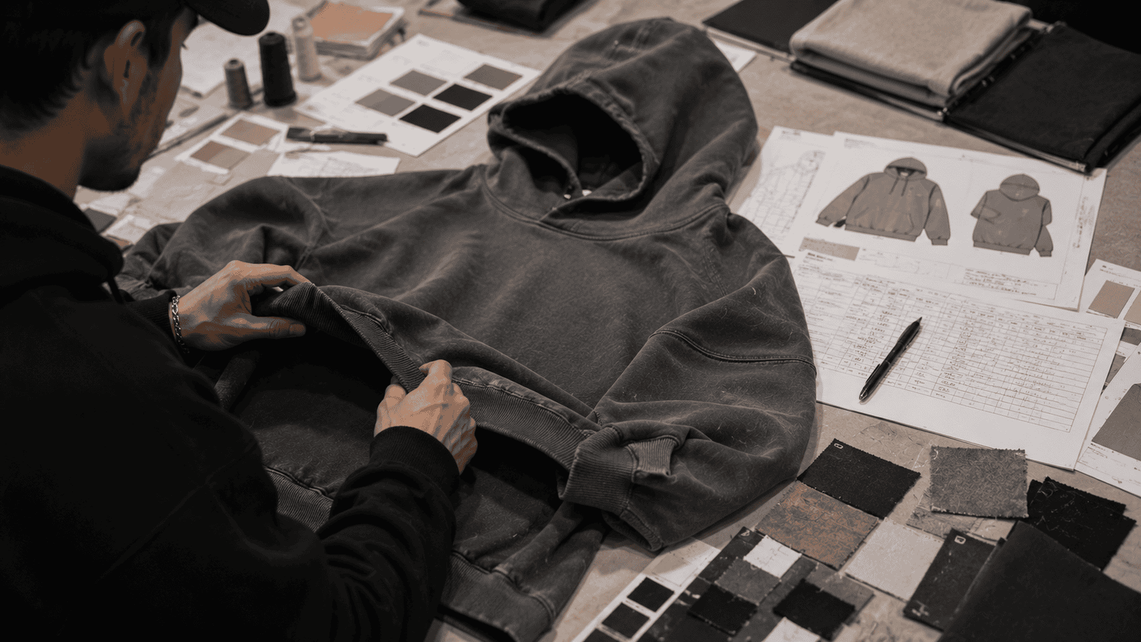 Streetwear hoodie sample development process, garment sample laid on table with tech pack, measurement sheet, fabric swatches and color cards, designer reviewing oversized fit details, professional apparel product development workflow, ultra realistic, high detail, no text
